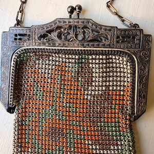 Op de afbeelding: Een vintage metalen mesh handtas met een bloemenpatroon in oranje, groen en cr&egrave;me. De tas heeft een sierlijk, donkerkleurig metalen frame met gedetailleerd krulwerk, een dubbele bolsluiting en een bijpassend metalen kettinghengsel.