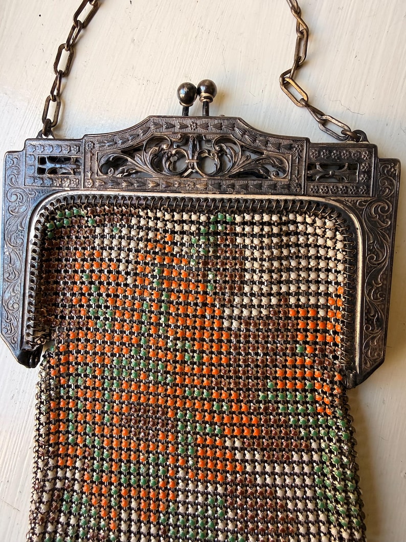 Op de afbeelding: Een vintage metalen mesh handtas met een bloemenpatroon in oranje, groen en cr&egrave;me. De tas heeft een sierlijk, donkerkleurig metalen frame met gedetailleerd krulwerk, een dubbele bolsluiting en een bijpassend metalen kettinghengsel.