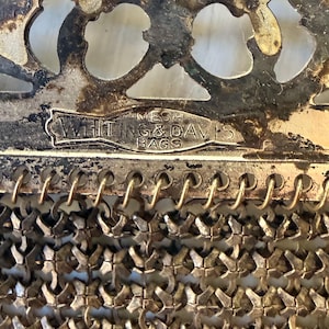 Op de afbeelding: Een goudkleurige metalen mesh tas met een sierlijk, uitgesneden filigraan frame. De metalen plaat is voorzien van de tekst Whiting & Davis Mesh Bags. De tas bestaat uit een fijn, in elkaar grijpend metalen gaaspatroon met een verweerde afwerking.
