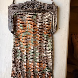 Op de afbeelding: Een vintage metalen mesh handtas met een bloemenpatroon in oranje, groen en bruin op een cr&egrave;mekleurige achtergrond. De tas heeft een sierlijk, gegraveerd zilverkleurig metalen frame met een knipsluiting en een dunne metalen ketting als hengsel.
