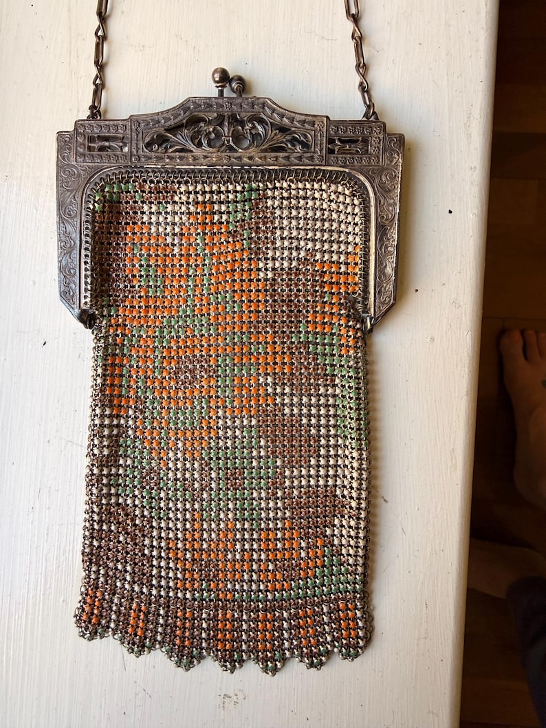 Op de afbeelding: Een vintage metalen mesh handtas met een bloemenpatroon in oranje, groen en bruin op een cr&egrave;mekleurige achtergrond. De tas heeft een sierlijk, gegraveerd zilverkleurig metalen frame met een knipsluiting en een dunne metalen ketting als hengsel.