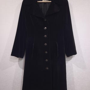 May include: A black velvet coat featuring a wide collar, long sleeves, and a front button closure with six dark buttons. The interior label displays the brand name Harella. The coat has a structured silhouette and reaches below the knee.
