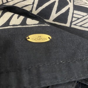 May include: Close-up of a black and cream-colored textile with a geometric pattern. The fabric features a repeating design of triangles and rectangles. A small, gold-colored oval tag with the text "JIM THOMPSON" is sewn onto the black fabric.