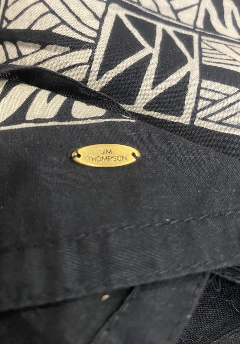 May include: Close-up of a black and cream-colored textile with a geometric pattern. The fabric features a repeating design of triangles and rectangles. A small, gold-colored oval tag with the text "JIM THOMPSON" is sewn onto the black fabric.