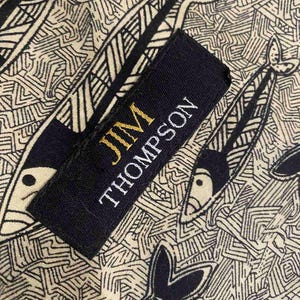 May include: Close-up of a fabric with a repeating fish pattern in black and cream. A black label with gold text reads "JIM THOMPSON". The fabric design features stylized fish and geometric shapes.