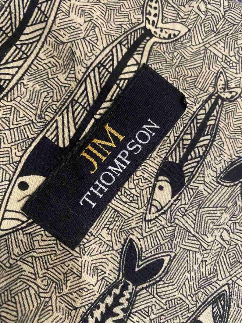 May include: Close-up of a fabric with a repeating fish pattern in black and cream. A black label with gold text reads "JIM THOMPSON". The fabric design features stylized fish and geometric shapes.