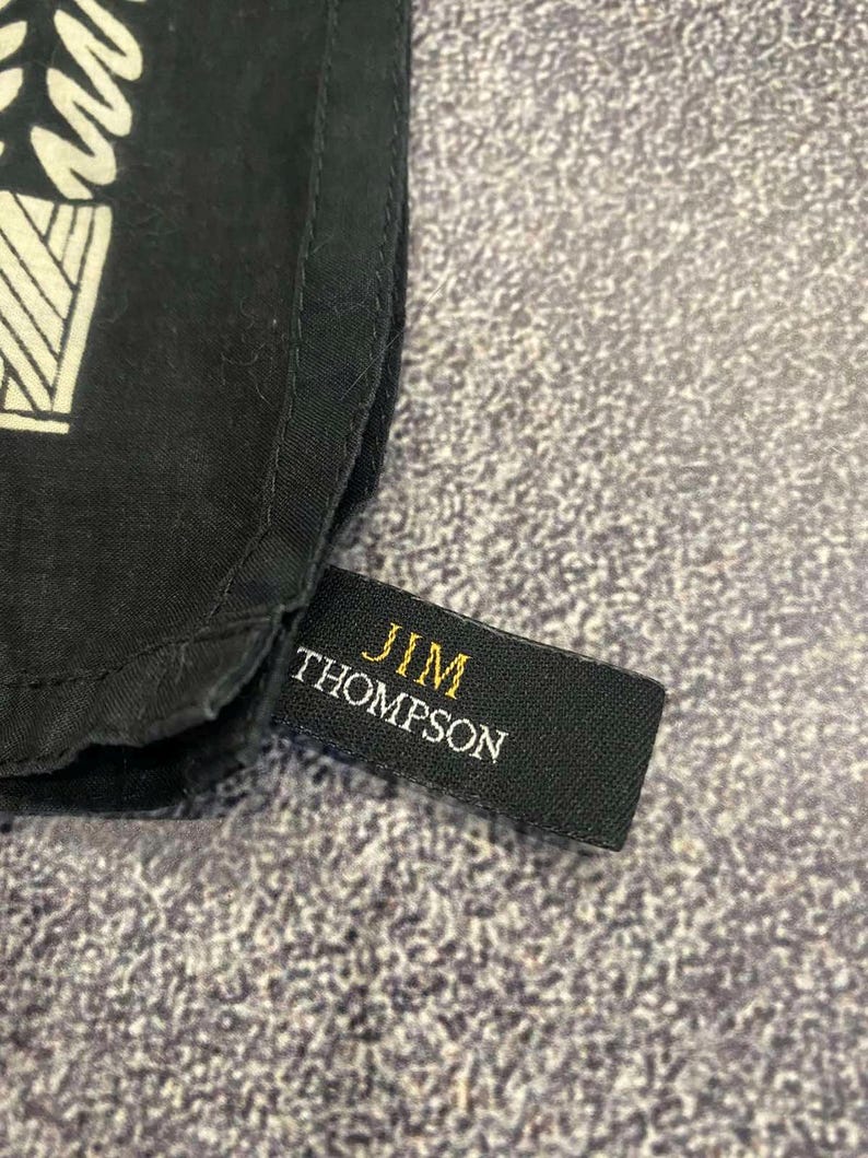 May include: Close-up of a black fabric with a white geometric design. A black tag with the gold text "JIM THOMPSON" is attached to the fabric. The fabric has a textured appearance.