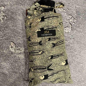 May include: A drawstring bag with a repeating pattern of black and white fish on a light green background. The bag has a black label with the text "JIM THOMPSON". The bag is made of a soft, patterned fabric.
