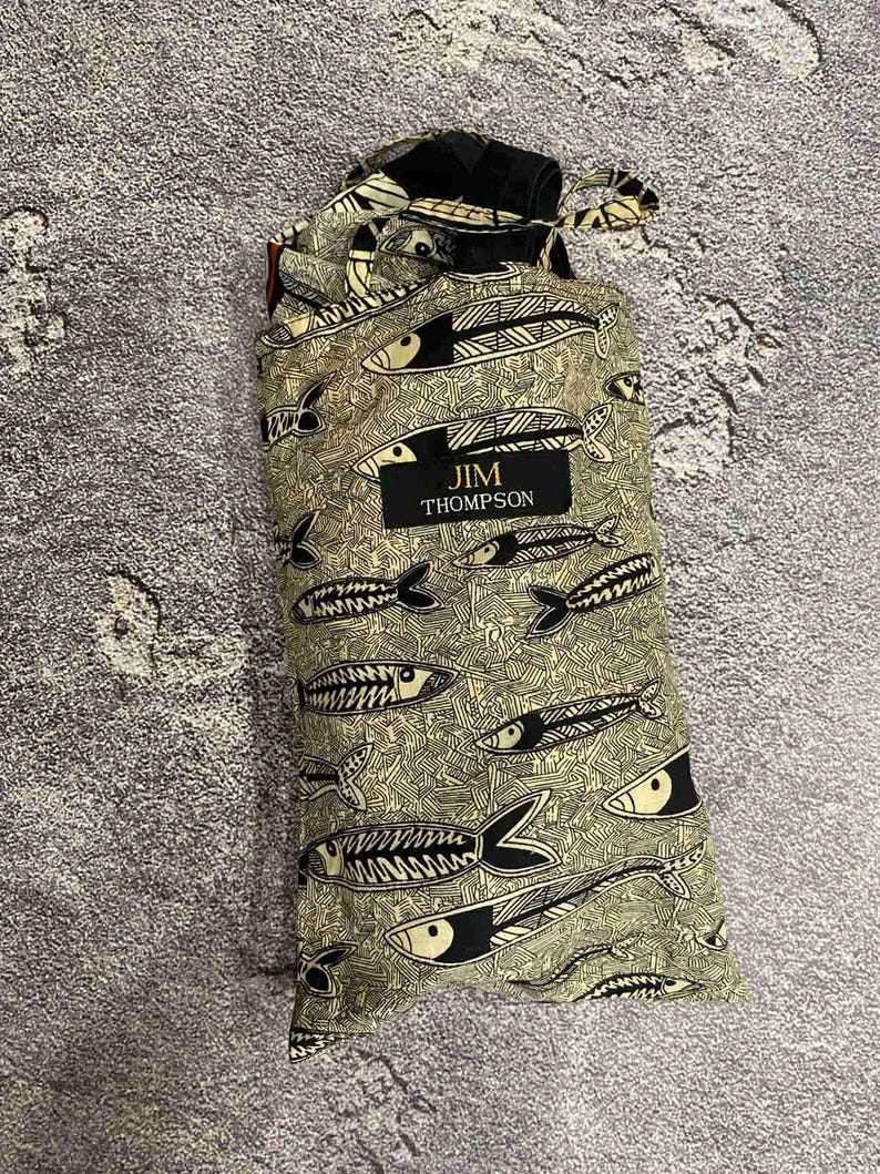 May include: A drawstring bag with a repeating pattern of black and white fish on a light green background. The bag has a black label with the text "JIM THOMPSON". The bag is made of a soft, patterned fabric.