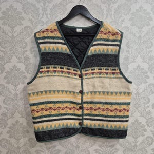 Op de afbeelding: Een mouwloos vest met een veelkleurig horizontaal patroon in tinten crème, antracietgrijs, geel, blauwgroen en bordeauxrood. Het kledingstuk heeft een V-hals, een knoopsluiting aan de voorzijde met vier bruine knopen en een zwarte gewatteerde voering.