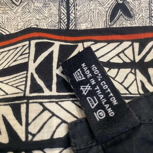 May include: Close-up of a black and white patterned fabric with a red stripe. The fabric has a geometric design and a tag that reads "100% Cotton Made in Thailand."