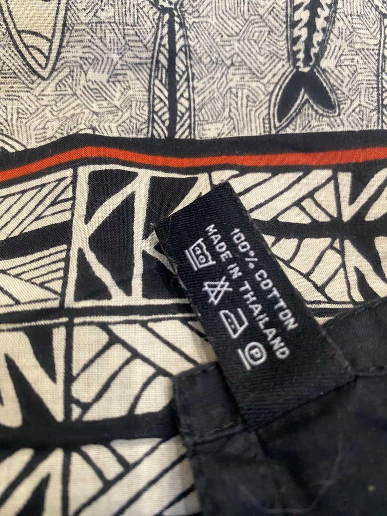 May include: Close-up of a black and white patterned fabric with a red stripe. The fabric has a geometric design and a tag that reads "100% Cotton Made in Thailand."