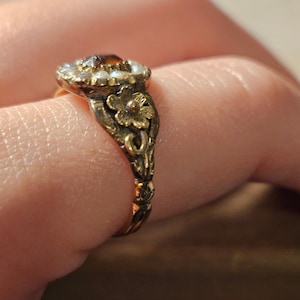 Könnte beinhalten: Ein goldener Vintage-Ring mit einem bernsteinfarbenen Mittelstein, umgeben von kleinen weißen Perlen. Band und Fassung des Rings sind mit floralen Mustern verziert, die einen antiken Stil widerspiegeln. Schmuck.
