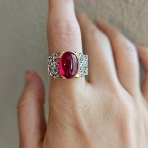 Op de afbeelding: Een ring met een grote, ovale, dieprode cabochon edelsteen gezet in geelgoud, geflankeerd door twee rechthoekige secties met kleine, heldere, rondgeslepen stenen in een zilverkleurige metalen band met een getrapt, geometrisch ontwerp.