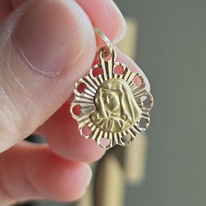 May include: A circular gold pendant featuring a central embossed profile of a figure wearing a head covering, surrounded by a sunburst-style border with cut-out details. The pendant includes a small top loop for attachment to a chain or cord.