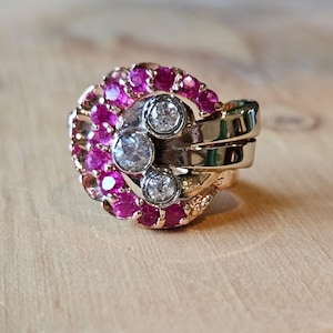 Puede incluir: Un anillo de oro con un engaste curvo en forma de media luna adornado con una hilera de rubíes de color rosa vibrante. El centro del anillo muestra tres diamantes blancos redondos y claros dispuestos en línea vertical sobre una banda de oro pulido.