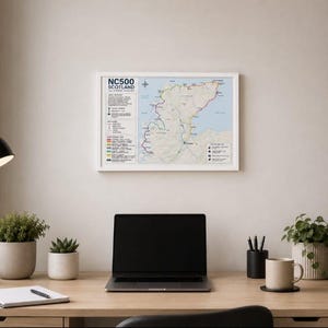 Può includere: Una mappa da parete incorniciata intitolata NC500 Scotland, che presenta un tracciato dettagliato del percorso, una rosa dei venti e una legenda con chiavi a colori. La mappa mostra il terreno geografico in toni beige e blu, in una cornice bianca.