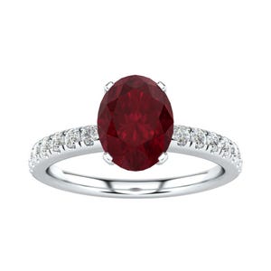 May include: A silver-toned ring featuring a large, oval-cut deep red gemstone set in the center, held by four prongs. The band is adorned with a series of small, round, clear white stones embedded along both sides of the shank, creating a sparkling effect.