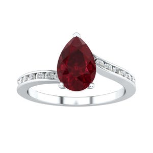 May include: A silver-toned ring featuring a large, pear-shaped deep red gemstone at the center. The band is split into two curved sections that bypass the central stone, each channel-set with a row of small, clear, round-cut white accent stones.