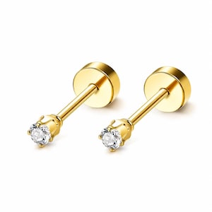 May include: A pair of gold-tone stud earrings featuring a clear, round-cut gemstone set in a four-prong basket setting. Each earring has a long, straight post and a flat, circular screw-back closure, providing a secure fit for the wearer.