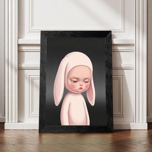 May include: A framed wall art print featuring a pale pink character with long, drooping rabbit ears and a somber expression. The character is centered against a solid dark gray background, enclosed in a simple black rectangular frame.