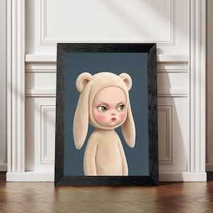 May include: A framed wall art print featuring a stylized character with a grumpy expression, wearing a beige hooded onesie with long, floppy rabbit-like ears. The character has large green eyes and a small red mouth, set against a solid dark blue background.