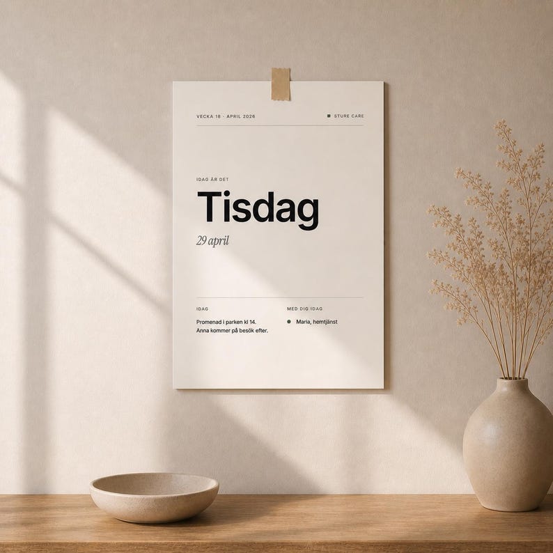 May include: A beige poster with the word "Tisdag" in large black font, along with the date "29 april". The poster is attached to a wall with a piece of tape. A vase with dried plants and a small bowl are in the foreground.