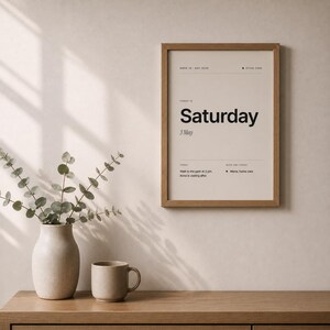 May include: A wooden-framed print displaying "Saturday" in a large black font, with the date "3 May". A beige vase with eucalyptus and a matching mug are on a wooden surface, creating a minimalist aesthetic.