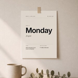 May include: A beige daily planner with the word "Monday" in large black font. The date "28 April" is below. The planner includes a to-do list and is taped to a wall. A beige mug and greenery are on a wooden surface.