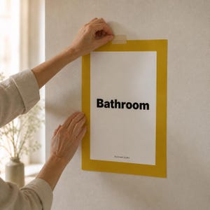 May include: A rectangular sign with a yellow border and the word "Bathroom" in bold black letters. The sign is being taped to a white wall. The sign is a decorative element for a bathroom.