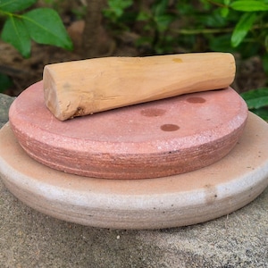 May include: A cylindrical light brown wooden pestle rests horizontally on top of a circular reddish-brown stone grinding slab, which is positioned on a larger, thicker, circular light beige stone base with a smooth, rounded edge.
