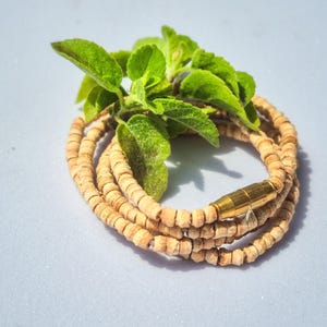 Puede incluir: Un collar de múltiples hilos compuesto por pequeñas cuentas cilíndricas de madera de color marrón claro. El collar presenta un cierre de barril de metal en tono dorado pulido y está dispuesto en una espiral circular, con hojas verdes.