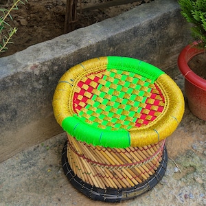 May include: A round stool featuring a woven seat with a vibrant pattern of bright green, gold, and red plastic strips. The circular frame is wrapped in gold and green cord, while the base consists of vertical wooden sticks bound by dark wire.