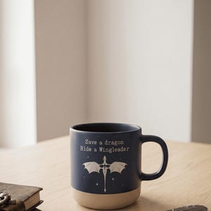 Può includere: Una tazza in ceramica blu navy con base color crema, con un drago e la scritta "Save a dragon Ride a Wingleader". Una penna decorativa e un diario rilegato in pelle sono su una superficie di legno.