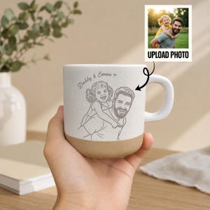 Peut inclure: Une tasse en céramique blanche avec une base beige, représentant un croquis d'un père et d'un enfant. La tasse porte les mots "Daddy & Emma" et une option de téléchargement de photo. La tasse est tenue dans une main.