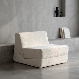 May include: A cream-coloured, modern armchair with a textured, plush fabric. The chair has a low profile with a wide seat and a slightly angled backrest. The design is minimalist and contemporary, suitable for a living room or lounge area.