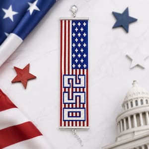 Op de afbeelding: Een patriottische armband met het ontwerp van de Amerikaanse vlag met rode en witte strepen, een blauw veld met witte sterren en de tekst "USA". De armband heeft een zilveren sluiting en ketting. De armband is ongeveer 18 cm lang.