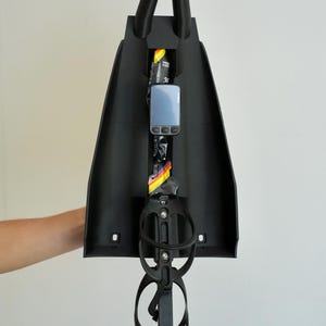 Puede incluir: Accesorio de bicicleta negro con dos asas verticales en la parte superior y un portabotellas en la parte inferior. Un pequeño dispositivo electrónico y un paquete de combustible son visibles en el interior.