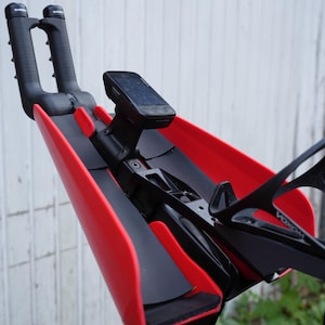 May include: A black and red bicycle handlebar attachment with a mounted device and water bottle cage. The attachment features a red aerodynamic design with black accents and a digital device mounted in the center.