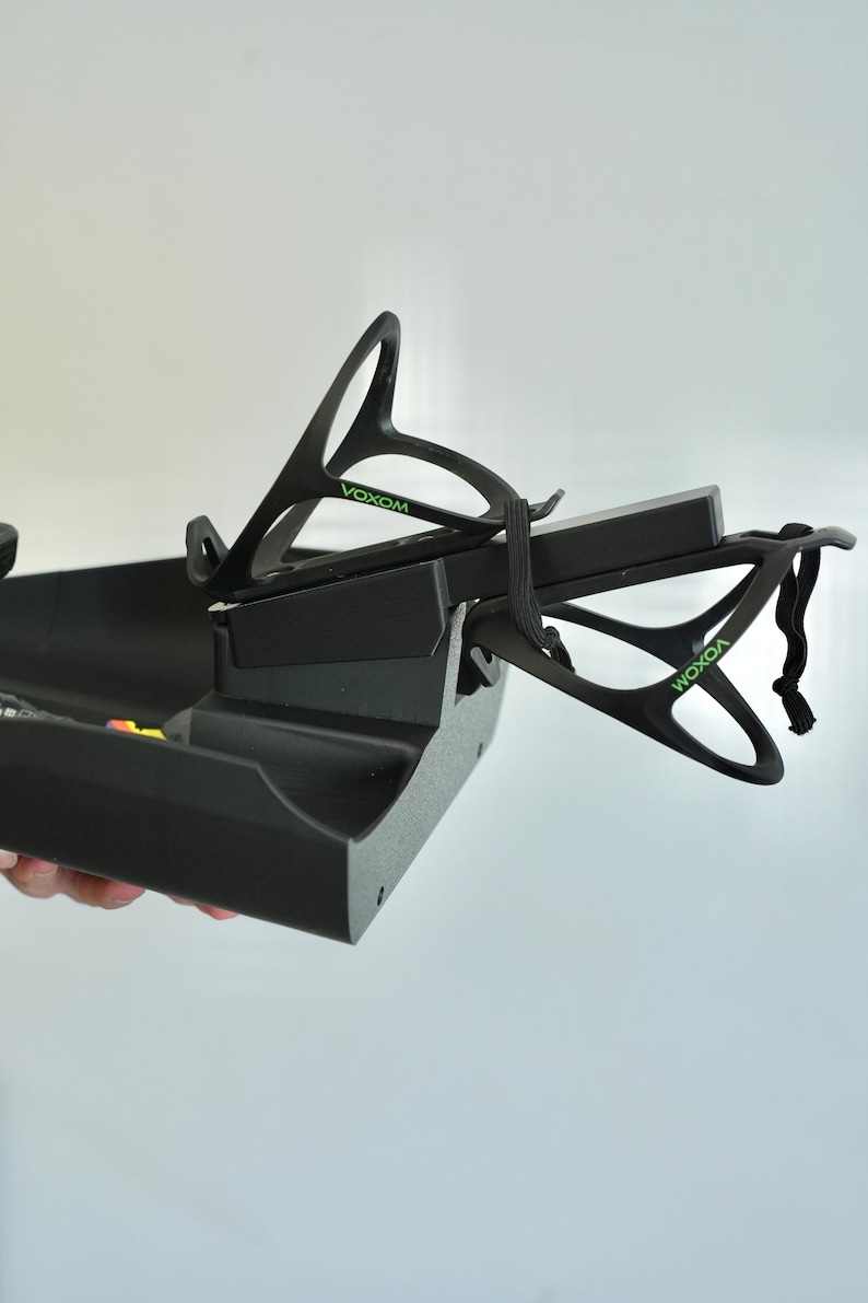 May include: Black plastic bicycle accessory with the word "VOXOM" in green. The accessory has a tray and two holders for water bottles. The holders are attached to the tray with a black elastic strap.
