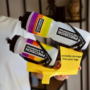 Könnte beinhalten: Eine leuchtend gelbe Kunststoffhalterung mit zwei transparenten Wasserflaschen mit schwarzen Verschlüssen. Jede Flasche hat ein regenbogenfarbenes Band und den Text "PRECISION FUEL & HYDRATION". Die gelbe Halterung zeigt den schwarzen Text "probably stronger than your legs".