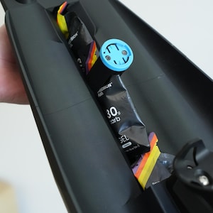 May include: Black sports equipment with a blue and black accessory. The equipment holds energy gel packets and a blue device. The gel packets are black with the text "PRECISION FUEL" and "30g carb".