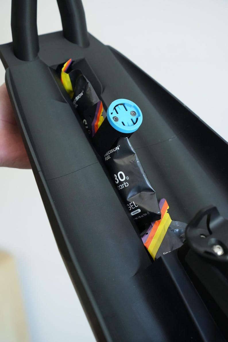 May include: Black sports equipment with a blue and black accessory. The equipment holds energy gel packets and a blue device. The gel packets are black with the text "PRECISION FUEL" and "30g carb".