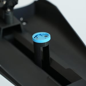 May include: A black plastic component with a light blue circular piece on top. The circular piece has a groove around the edge. The component is set against a white background.