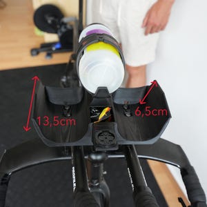 Pu&ograve; includere: Un supporto per idratazione nero per prolunghe da bicicletta con portaborraccia. Il supporto presenta un alloggiamento centrale per borraccia e supporti laterali. Frecce rosse indicano dimensioni di 13,5 cm per l'altezza laterale e 6,5 cm per la larghezza interna. Un supporto per ciclocomputer &egrave; integrato nella parte anteriore.