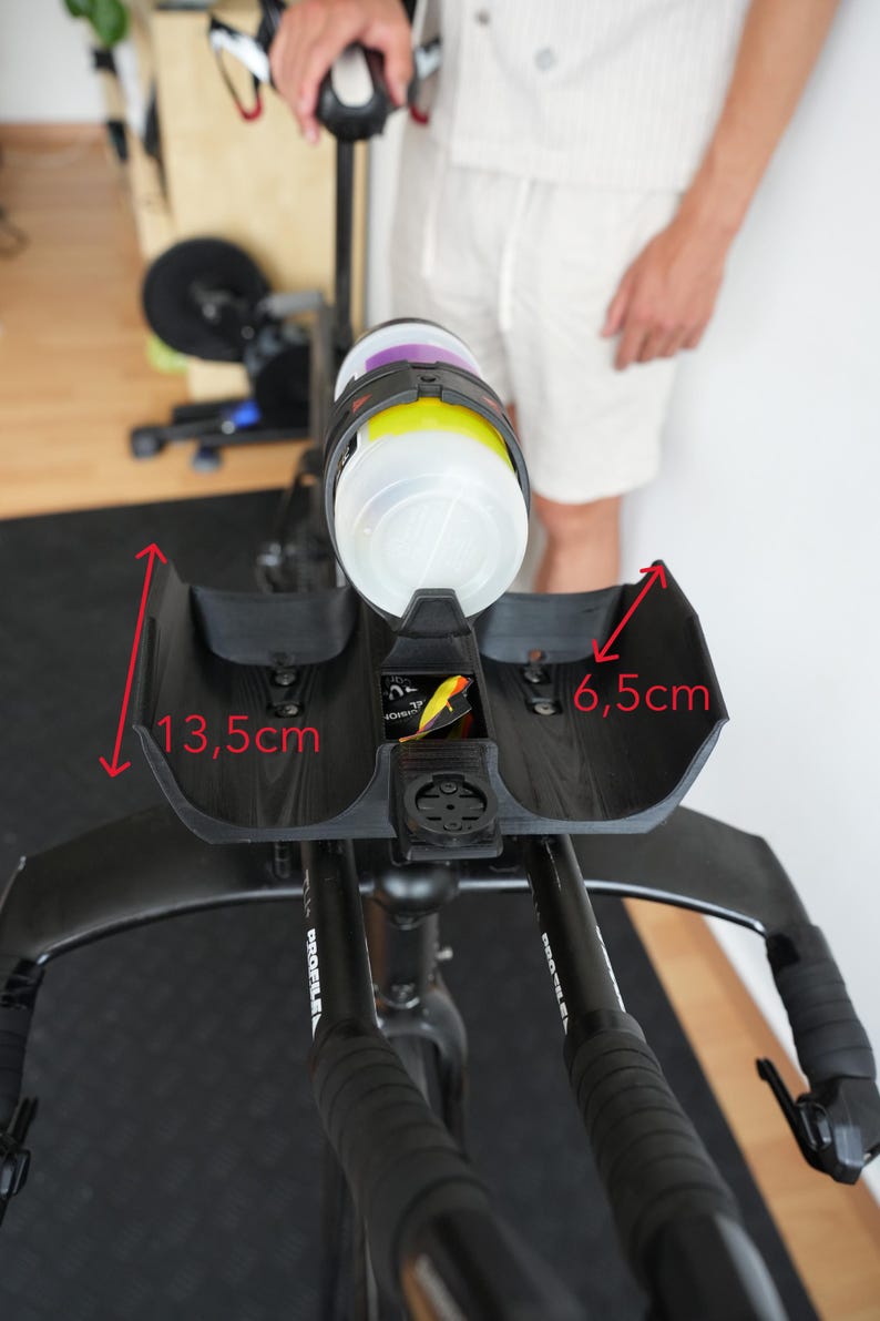 Pu&ograve; includere: Un supporto per idratazione nero per prolunghe da bicicletta con portaborraccia. Il supporto presenta un alloggiamento centrale per borraccia e supporti laterali. Frecce rosse indicano dimensioni di 13,5 cm per l'altezza laterale e 6,5 cm per la larghezza interna. Un supporto per ciclocomputer &egrave; integrato nella parte anteriore.
