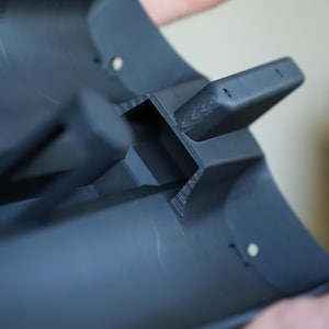 May include: A dark gray, curved plastic component with a rectangular protrusion and two small holes. The object is held by a person, showcasing its shape and design. The component appears to be 3D-printed.