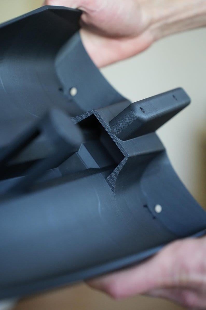 May include: A dark gray, curved plastic component with a rectangular protrusion and two small holes. The object is held by a person, showcasing its shape and design. The component appears to be 3D-printed.