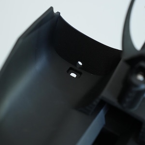 May include: Close-up of a black plastic component with a textured surface and a small, round hole. The component has a curved shape and appears to be part of a larger assembly. A small, rectangular opening is also visible.