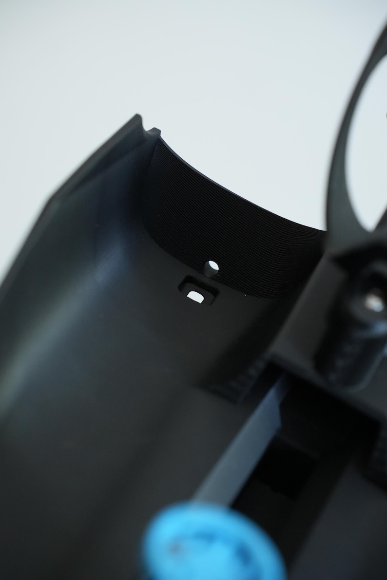 May include: Close-up of a black plastic component with a textured surface and a small, round hole. The component has a curved shape and appears to be part of a larger assembly. A small, rectangular opening is also visible.